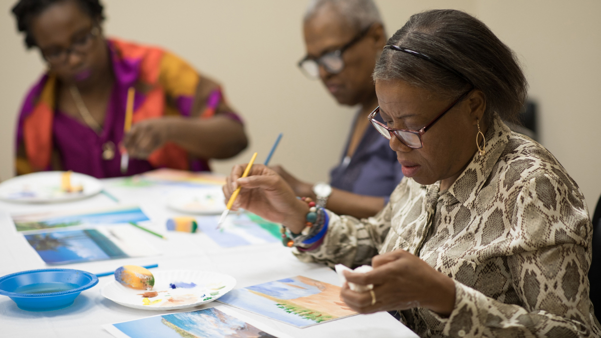 Three participants seated at a table work on landscape paintings.