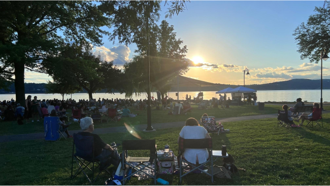 The sun sets behind hills framing a lake. Groups of people listen to the jazz concert as the sunlight filters through the trees over them.