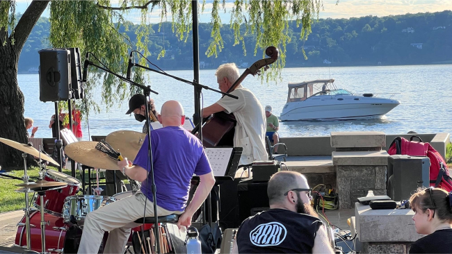 A small jazz ensemble plays in the park as a motor boat cruises along the lake behind them.