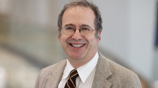 Dr. Ronald C. Samuels, MD, MPH. He smiles toward the camera, wearing a white shirt, maroon tie,  tan jacket and glasses.