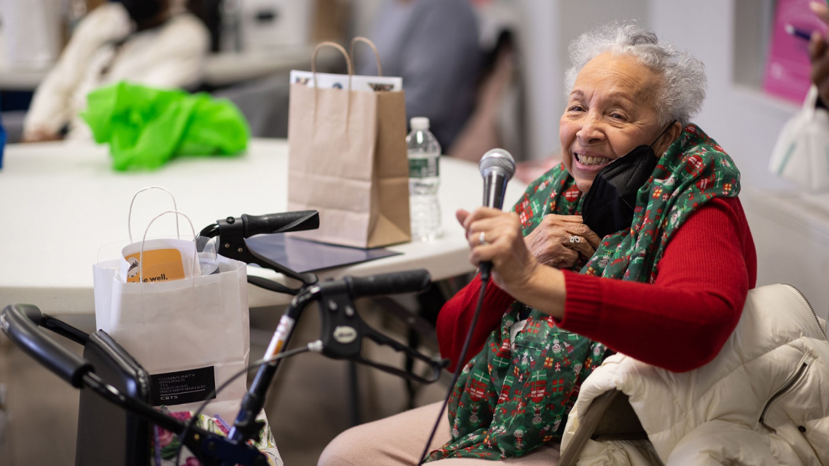 An older woman holding a microphone smiles toward the camera.