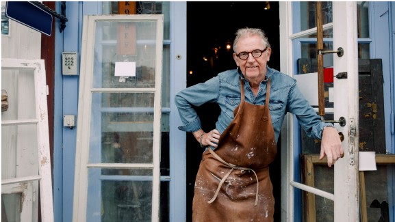 A man in a leather apron stands at the door of his workshop.