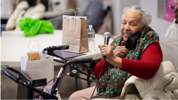 A woman in a chair holds a microphone and smiles toward the camera.