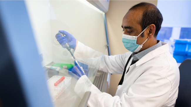 A gloved and masked scientist works with a pipette.