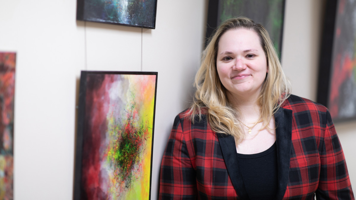 A woman in a red plaid shirt smiles, standing next to a wall covered in framed art.