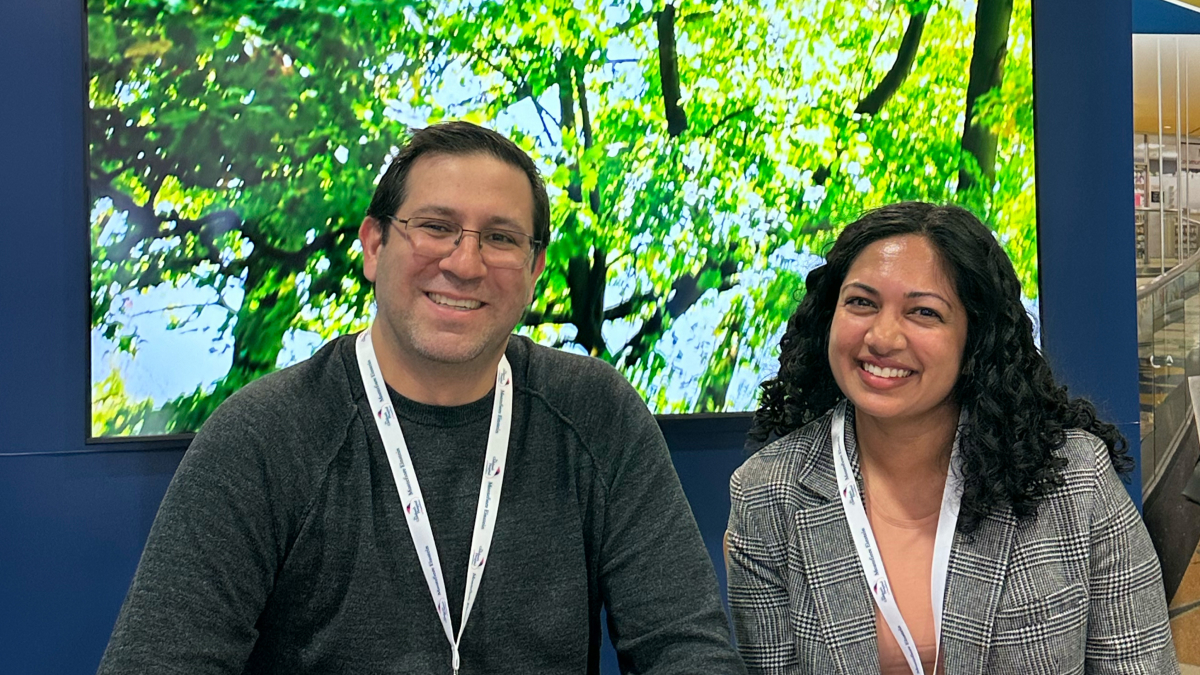 Two smiling participants stand in front of a woodland display on a monitor behind them.