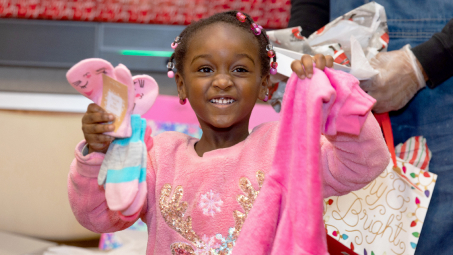 A young girl dressed in pink smiles widely, holding up presents.