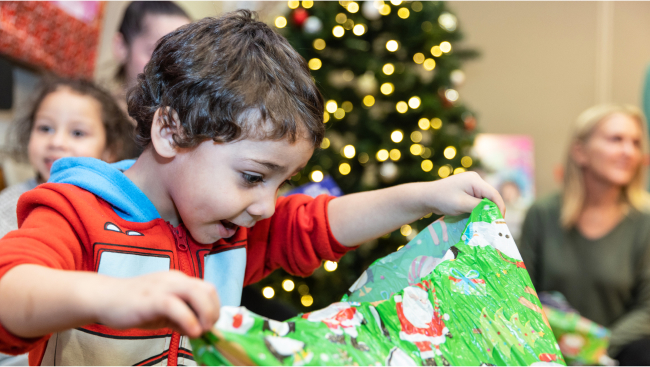 An excited child opens a gift wrapped in green paper.  Smiling faces and twinkling decorations are in the background.