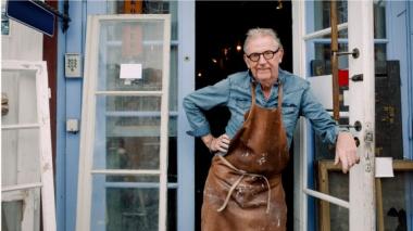 A man in a leather apron stands at the door of his workshop.