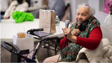 A woman in a chair holds a microphone and smiles toward the camera.