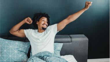 A young man stretches in bed with a smile on his face.