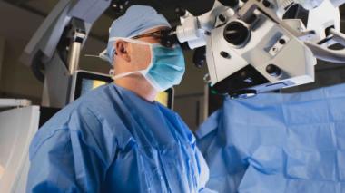 A surgeon dressed in blue scrubs works with a microscope guided tool.